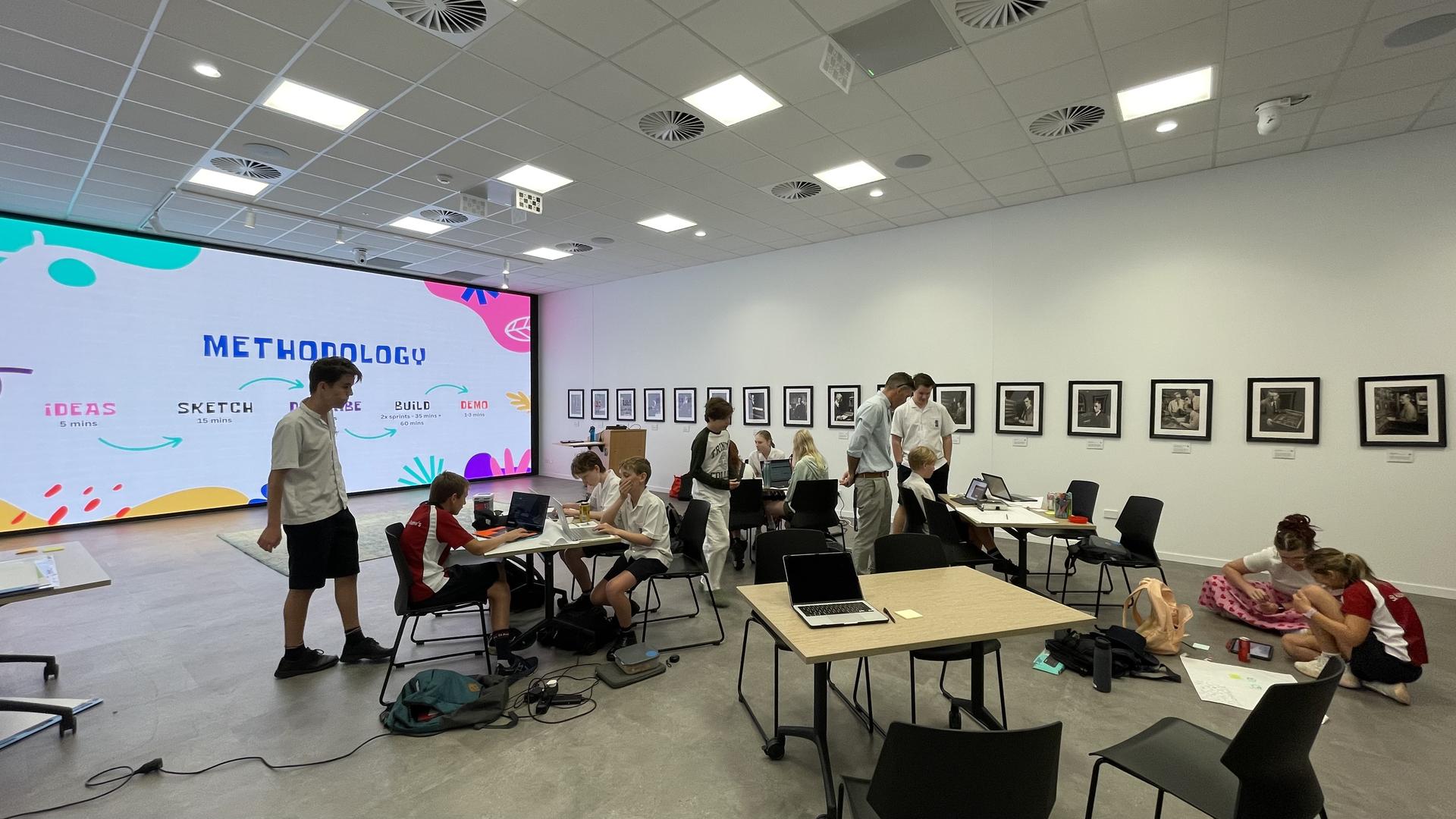 Wide view of the Hackathon room: students at tables with laptops, a large screen displaying the methodology (IDEAS, SKETCH, DESIGN, BUILD, DEMO), framed photographs along one wall.